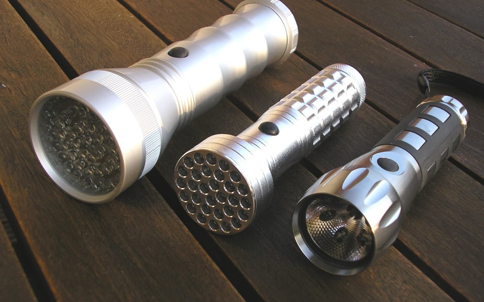 Three aluminium LED torches of different sizes lying side by side on dark wooden decking in daylight