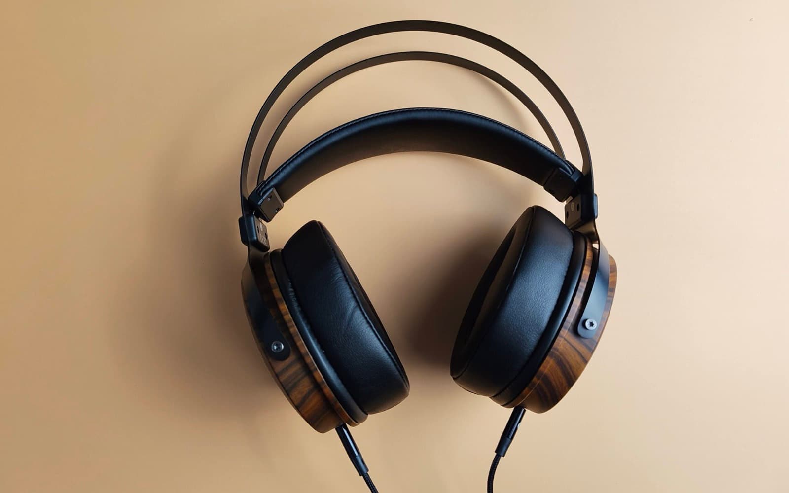 Wooden-cup over-ear headphones with black leather pads and braided cables on a plain warm background