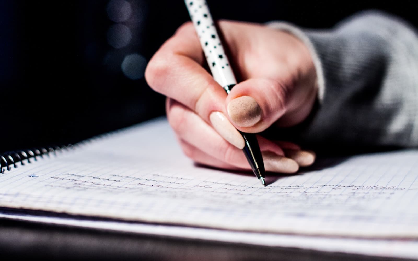 A hand writing with a ballpoint pen across the ruled pages of an open spiral-bound notebook