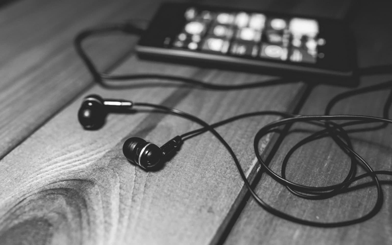 Black-and-white close-up of wired in-ear earphones and their looped cable on a wooden table, a phone behind