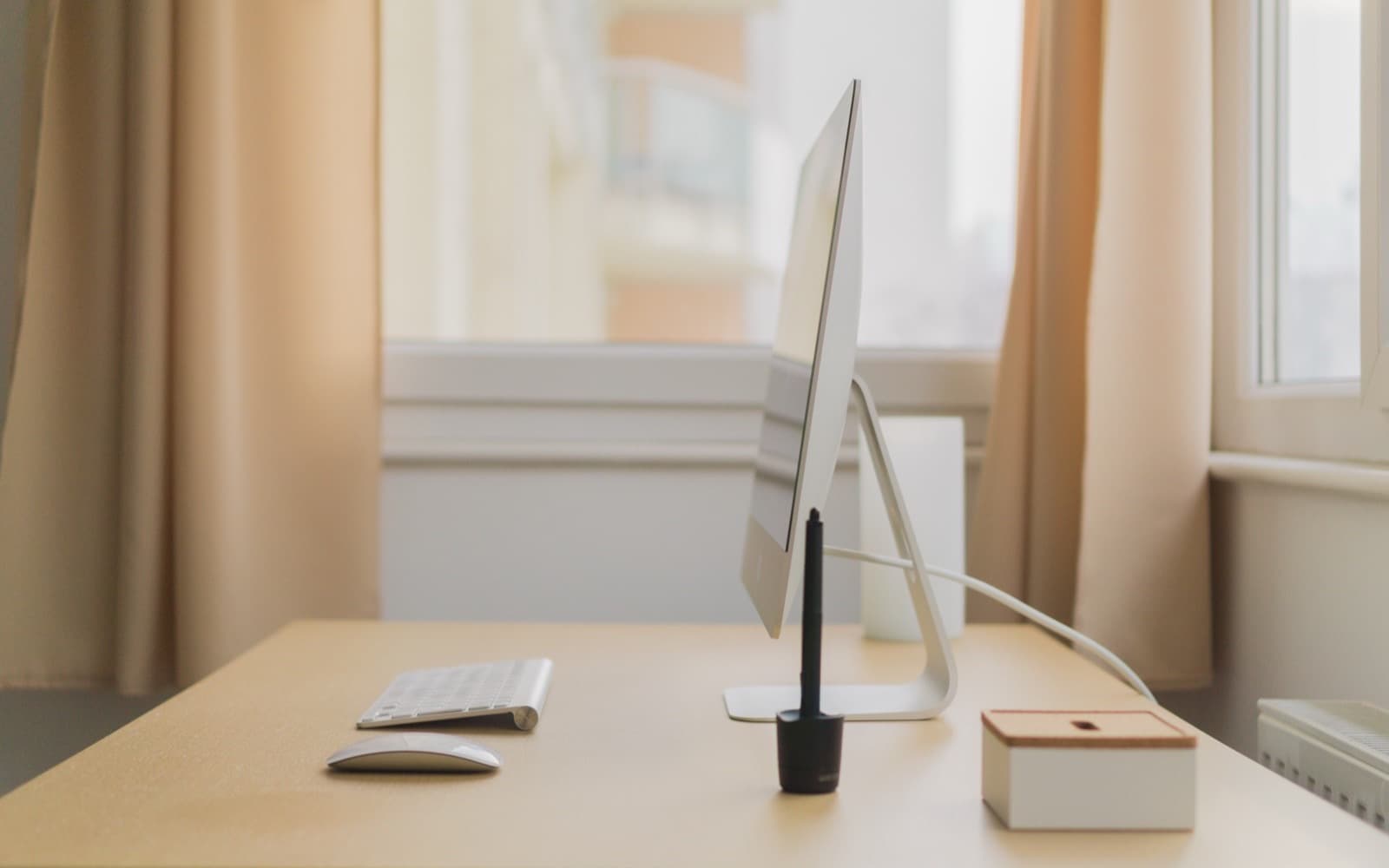 Side view of a slim monitor on its stand at a pale wooden desk, keyboard and mouse beside it, bright window behind
