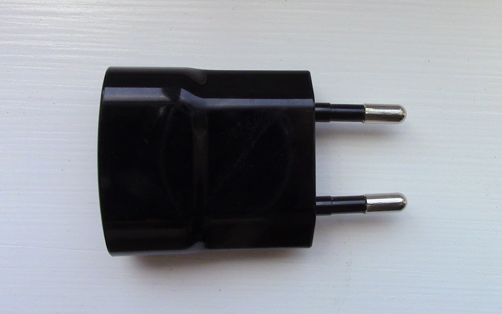 A black two-pin USB wall charger lying on a pale grey surface, its metal pins pointing right