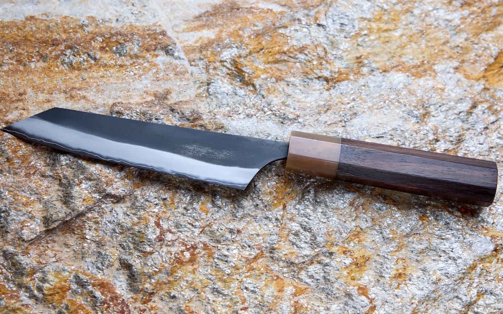 A forged Japanese kitchen knife with a dark hammered blade and octagonal wood handle, resting on sunlit stone