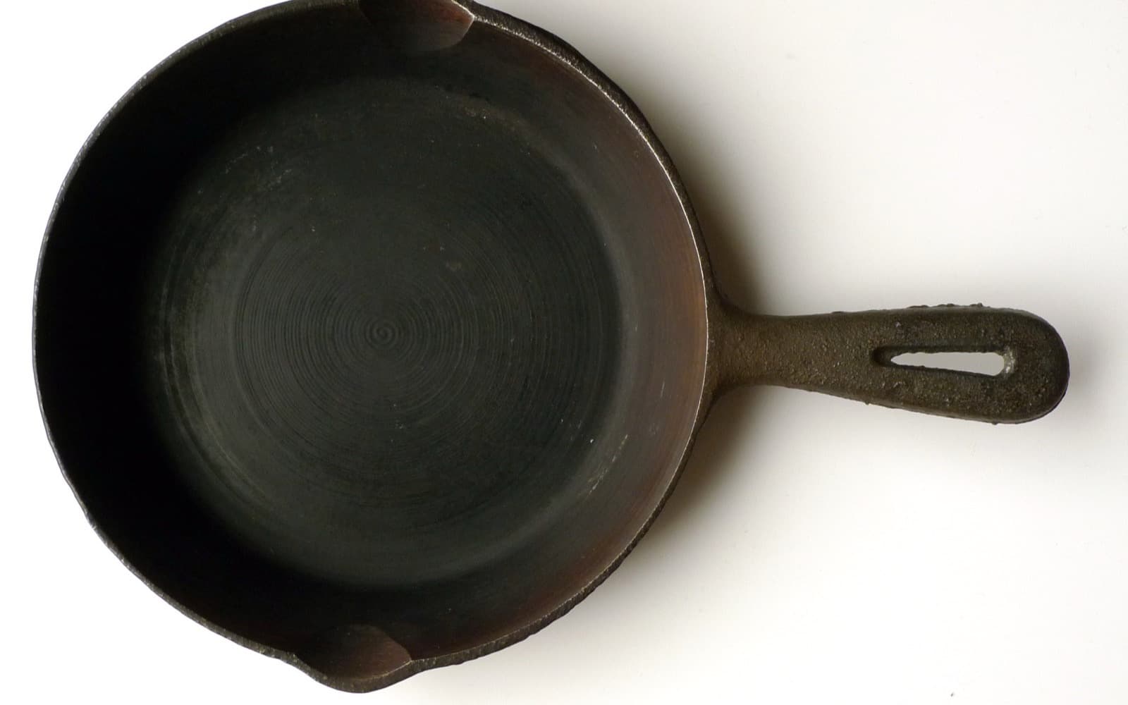 A bare cast iron skillet from above on a pale surface, spiral machining rings in the base and a slotted handle