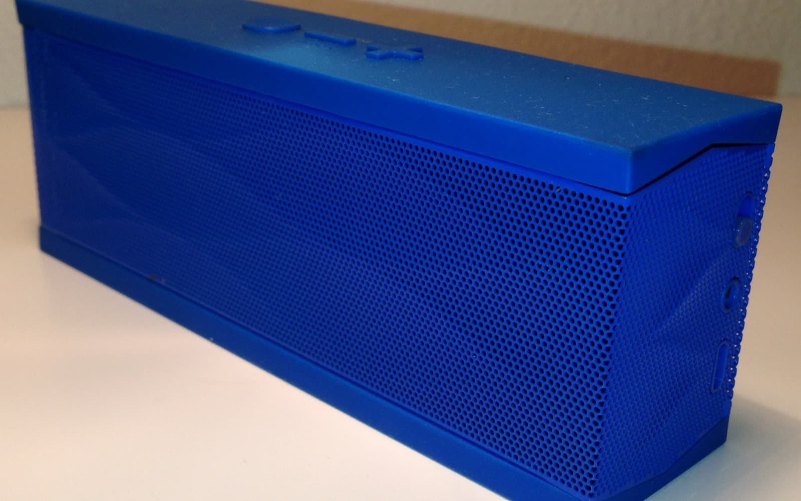 A small blue rectangular portable speaker with a perforated grille and raised playback buttons on top, on a white surface