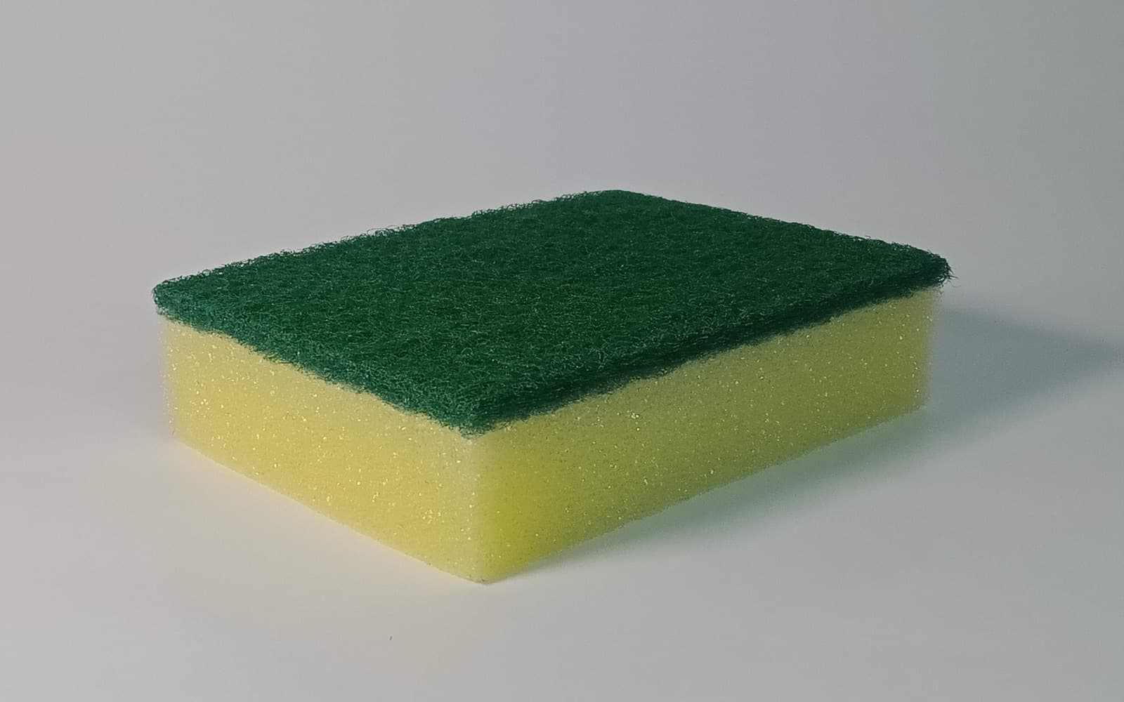 A yellow foam sponge topped with a green scouring pad, alone on a plain pale grey background