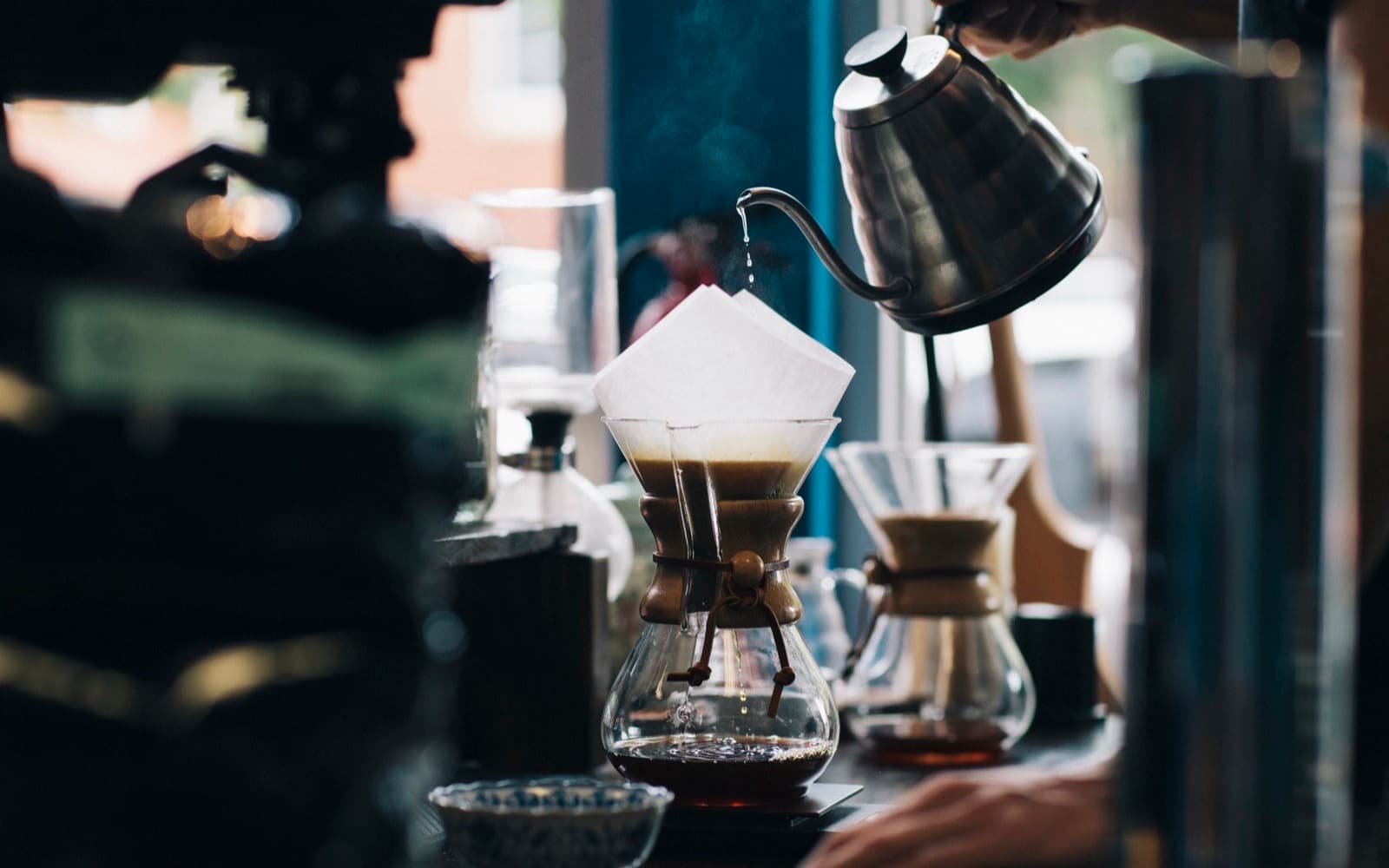 A gooseneck kettle pouring hot water into a glass pour-over brewer with a paper filter on a cafe counter