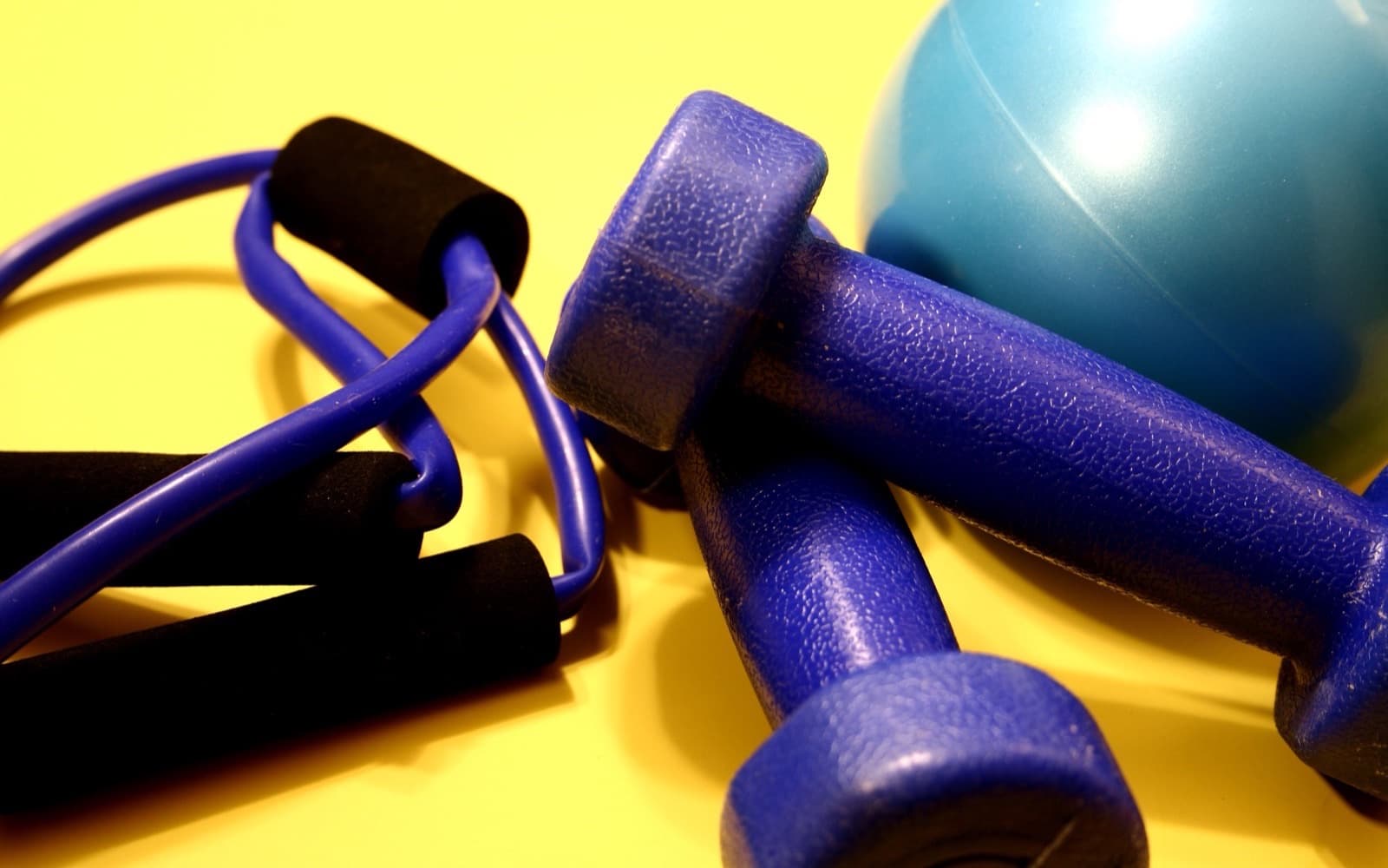 Two blue vinyl dumbbells, a resistance tube with foam handles and a teal exercise ball on a bright yellow surface