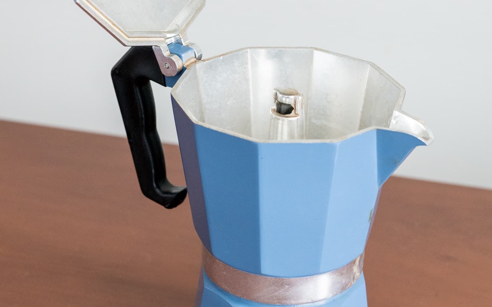 Pale blue aluminium moka pot on a wooden table, lid open, showing the empty upper chamber and its central spout