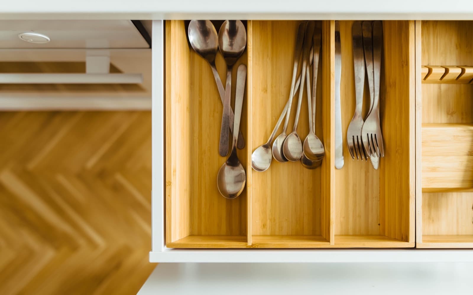 Steel spoons, ladles and forks sorted into the bamboo compartments of an open white kitchen drawer