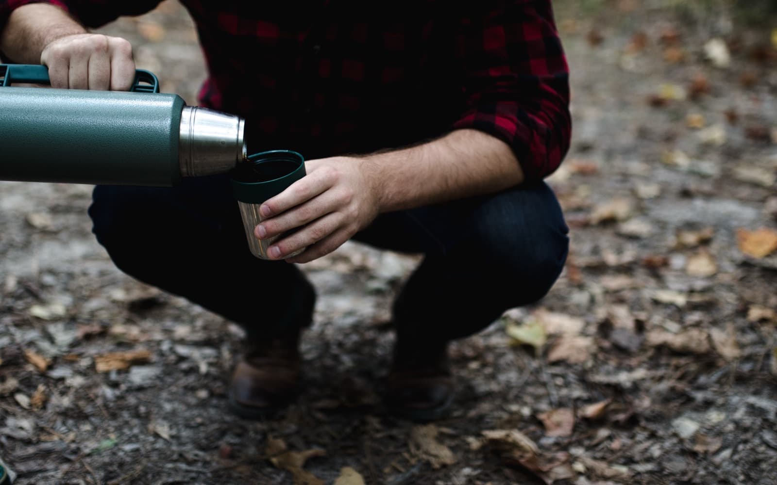 A green insulated flask being poured into its steel cup lid by someone crouching on a leafy forest floor