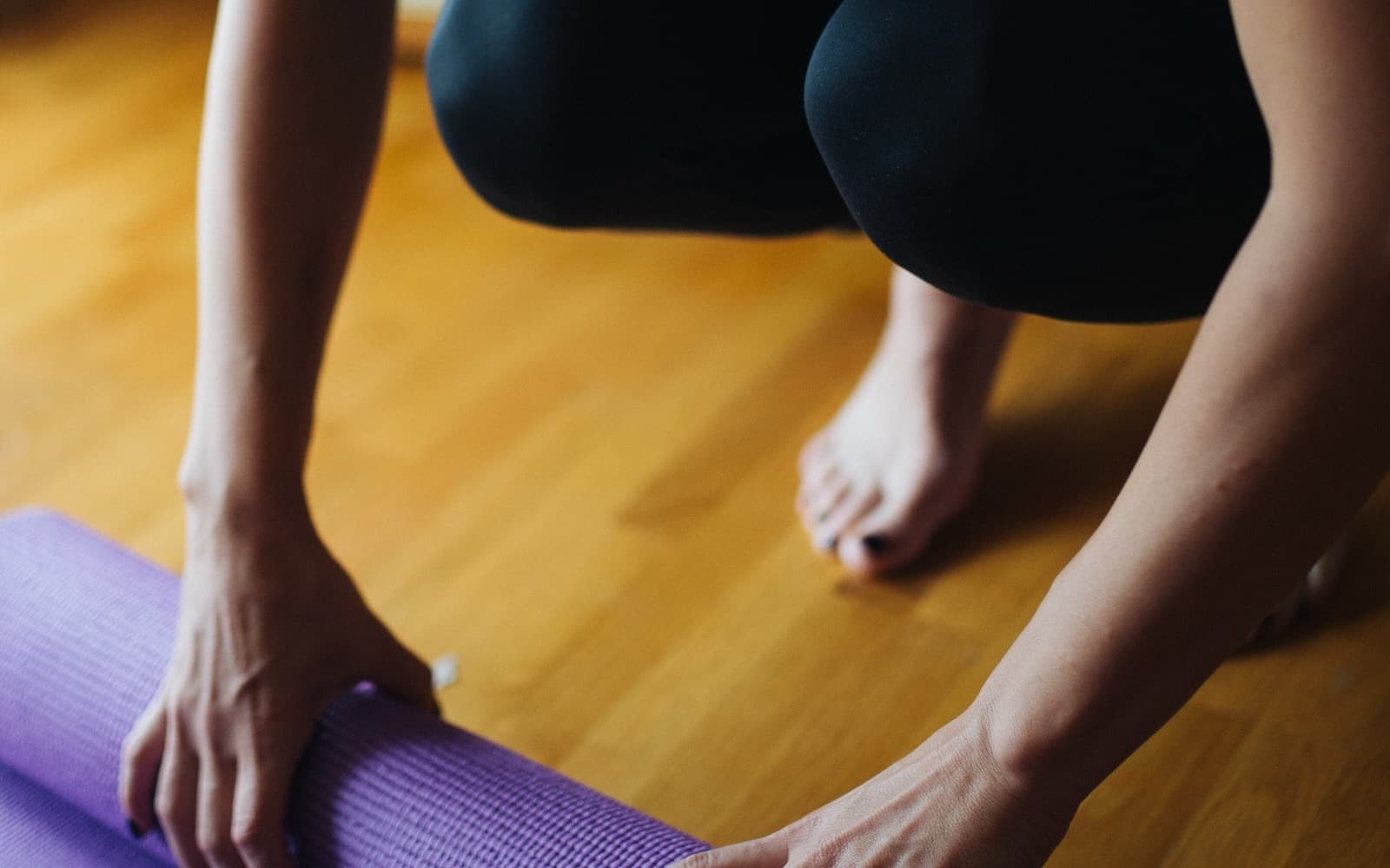 Hands rolling out a purple mat across a sunlit wooden floor, bare feet alongside